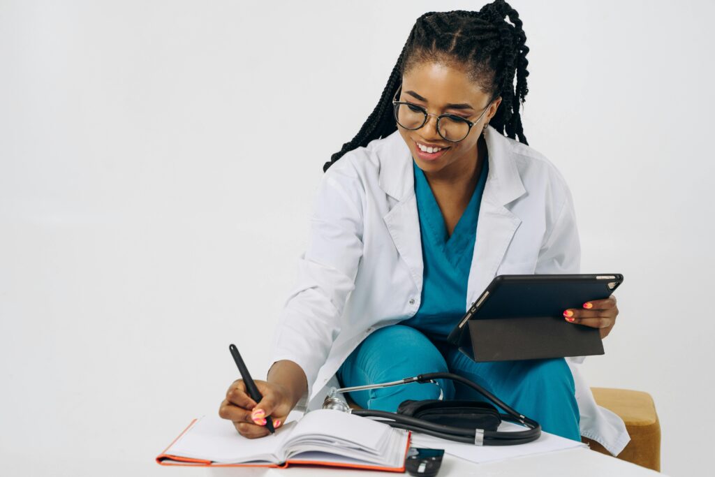 Smiling female doctor in scrubs and lab coat writing notes, holding a tablet.